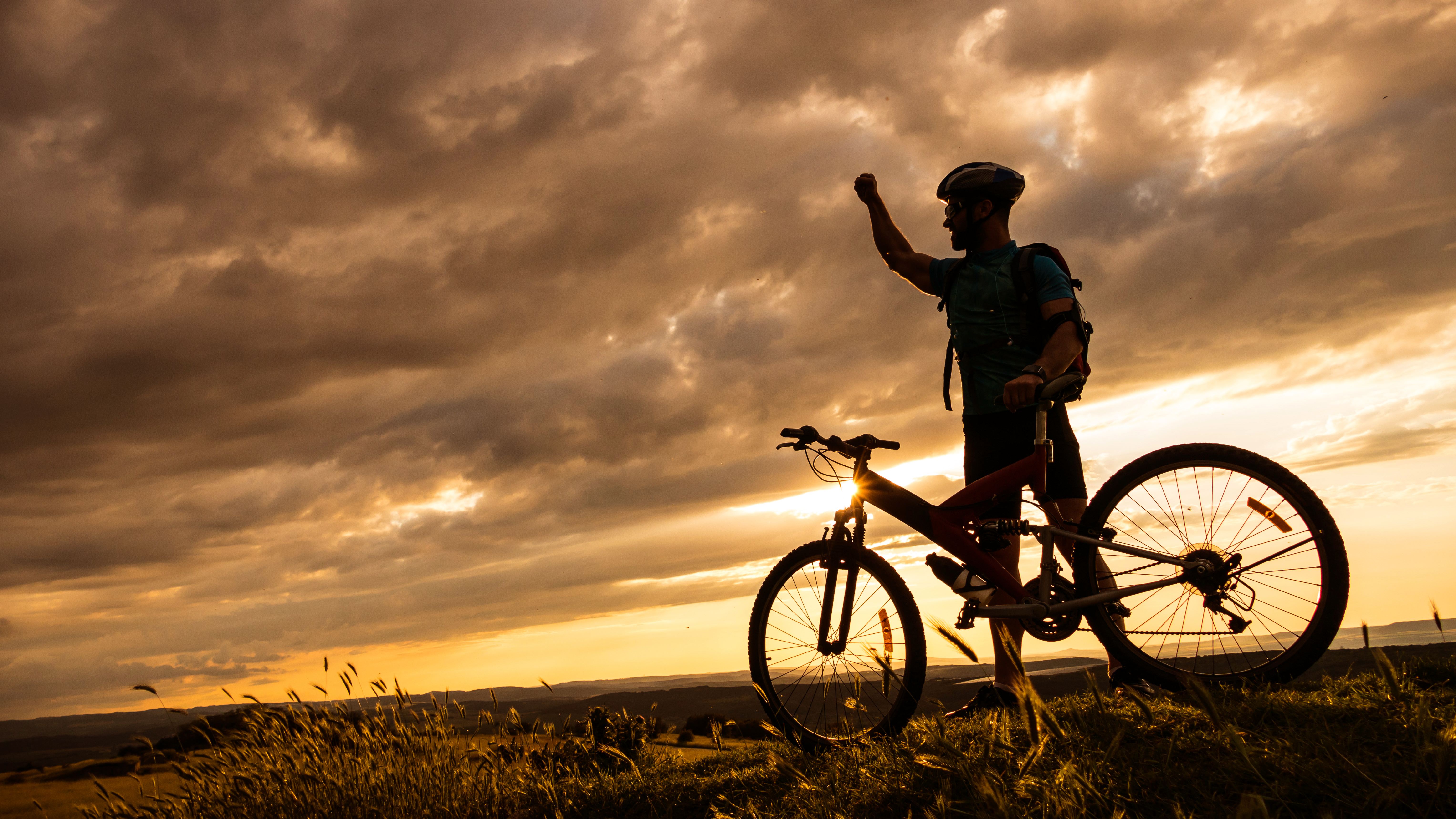 Silhouette of mountainbiker standing on top of the mountain with one hand raised. With helmet and backpack, arm band and headset. Sunbeam and clouds on background.
